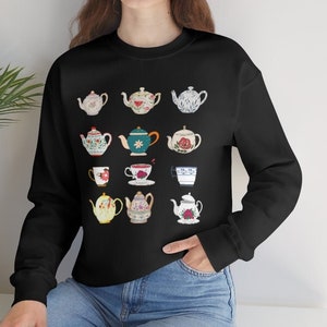 May include: Black sweatshirt featuring a collection of illustrated teapots in various colors and designs. The teapots are arranged in a grid pattern. The person is wearing blue jeans.