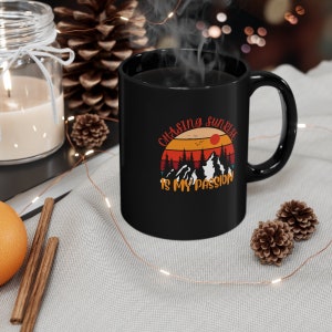 May include: Black ceramic mug with the text "Chasing Sunrise is My Passion" in a retro design. The design features a sunset, mountains, and trees in shades of orange, red, and white. The mug has a black handle and interior.