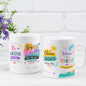 May include: Two white ceramic mugs featuring colourful motivational text and graphics. The left mug displays "Be GRATEFUL for today and every day" and "Stay FOCUSED CHASE YOUR GOALS not distractions". The right mug shows "Choose GROWTH every day" and "Believe in YOURSELF YOU CAN AND YOU WILL".