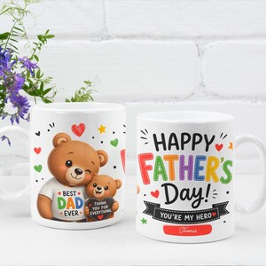 May include: Two white ceramic mugs featuring colorful graphics. One mug shows a large brown bear holding a smaller bear with the text BEST DAD EVER and THANK YOU FOR EVERYTHING. The other mug displays HAPPY FATHER'S DAY! YOU'RE MY HERO and Thomas.