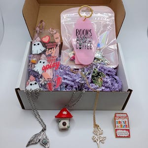 May include: A pink cardboard box filled with a variety of book-themed gifts, including a keychain that says "Books and Coffee", a ghost-shaped bookmark, a mushroom house charm, a rose charm, and a book fair pin.