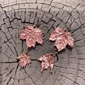 May include: A pair of copper earrings featuring a dangling maple leaf design. The earrings are made of a hammered copper metal and have a rose gold finish.