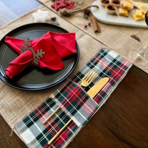 Puede incluir: Una servilleta roja con un anillo de servilleta de madera en forma de reno se encuentra sobre un plato negro. Un mantel individual de cuadros con un tenedor y un cuchillo dorados se encuentra sobre un camino de mesa de arpillera.