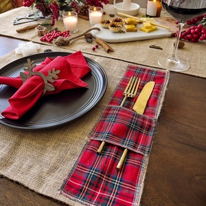 Puede incluir: Un mantel individual de cuadros rojos con un bolsillo para cubiertos. El mantel individual está sobre un camino de mesa de arpillera. Un plato negro con una servilleta roja y un anillo de servilleta de madera en forma de reno está a la izquierda. Un vaso de vino tinto está a la derecha.