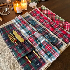 May include: Set of five plaid placemats with a burlap backing. The placemats are layered on top of each other, with the top layer featuring a green, red, and black plaid pattern. A gold fork and knife are placed in a pocket on the placemat.