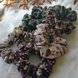May include: A set of six floral patterned scrunchies in various colors, including green, brown, beige, and purple. The scrunchies are made of fabric and have a gathered, ruffled look. Each scrunchie has a small tag attached with the word "Billy" written on it.
