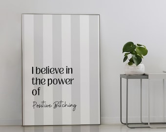 May include: A framed print with vertical gray and white stripes displays the text "I believe in the power of Positive Bitching." A small potted plant, books, and a lamp sit on two side tables. A striped rug is on the floor.
