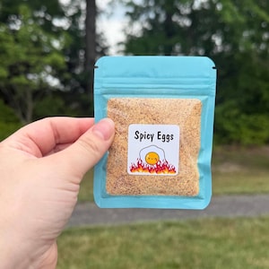 May include: A small, light blue resealable pouch containing a tan, granular seasoning mix. A square white label on the front features the text "Spicy Eggs" above a graphic of a fried egg with a smiling face surrounded by stylised red flames.