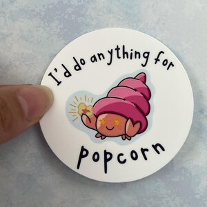 May include: A circular sticker with a cartoon crab illustration. The crab has a pink shell and star eyes, holding popcorn. The text "I'd do anything for popcorn" is written around the crab in black.