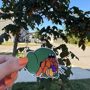May include: A colorful sticker featuring a cartoon hermit crab with an orange body, green shell, and a pumpkin. The sticker is held in front of a tree with green leaves and a blue sky in the background.