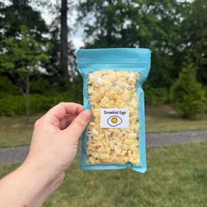 May include: A light blue resealable pouch filled with yellow popcorn. A white rectangular label on the front features the text Scrambled Eggs in black font above a simple graphic illustration of a fried egg with a yellow yolk and white edges.