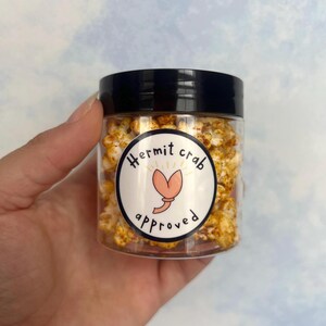 May include: A clear plastic jar filled with popcorn, topped with a black lid. The jar has a white label with the words "Hermit crab approved" and a cartoon crab illustration. The popcorn appears to be seasoned with a yellow spice.