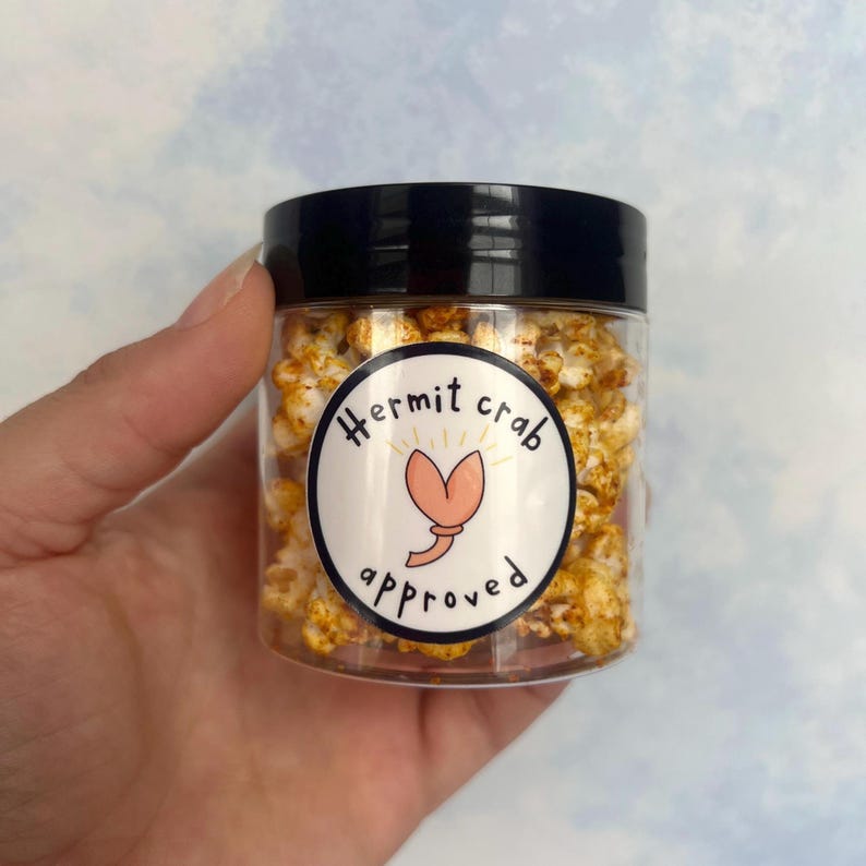May include: A clear plastic jar filled with popcorn, topped with a black lid. The jar has a white label with the words "Hermit crab approved" and a cartoon crab illustration. The popcorn appears to be seasoned with a yellow spice.