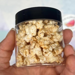 May include: A clear plastic jar filled with seasoned popcorn. The popcorn is a light beige colour with darker seasoning flecks. The jar has a black lid. The image is a close-up shot of the jar.