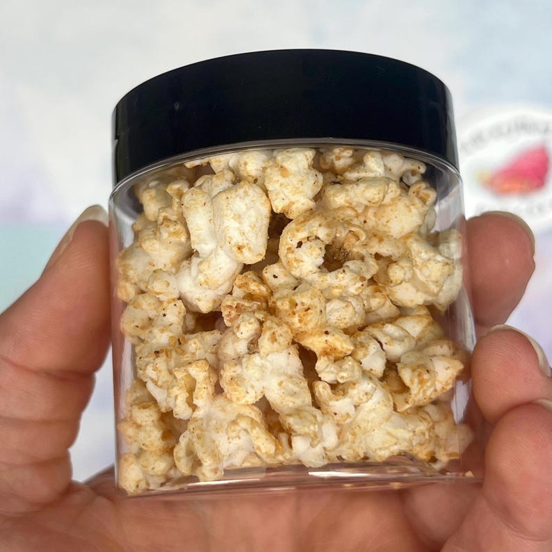 May include: A clear plastic jar filled with seasoned popcorn. The popcorn is a light beige colour with darker seasoning flecks. The jar has a black lid. The image is a close-up shot of the jar.