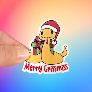 May include: A yellow snake wearing a red Santa hat holds a green and red gift box. The text "Merry Crissmiss" is written below the snake.