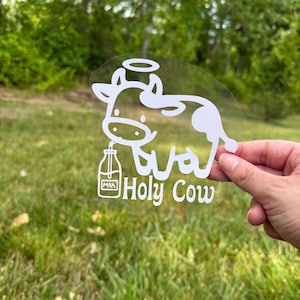 May include: A white vinyl decal featuring a cartoon cow with a halo above its head, drinking from a glass milk bottle. The text "Holy Cow" is printed in a stylised font below the cow, and the word "MILK" is visible on the bottle label.