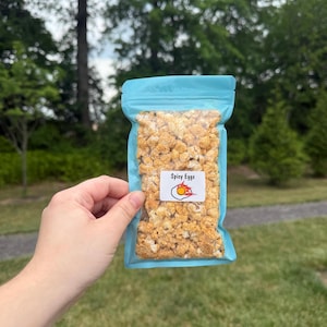May include: A light blue resealable plastic pouch filled with orange-dusted popcorn-like snacks. A white rectangular label on the front features the text Spicy Eggs and a graphic illustration of a fried egg with flames around the yolk.