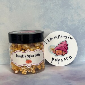 May include: A clear glass jar filled with pumpkin spice popcorn. The jar has a black lid and a white label that reads "Pumpkin Spice Latte". A white sticker with a cartoon crab and the text "I'd do anything for popcorn" is in the background.