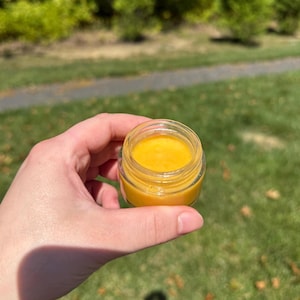 May include: A small, clear glass jar filled with a smooth, vibrant yellow substance. The container has a wide, circular opening and a thick, transparent base, showcasing the bright, uniform texture of the contents inside the vessel.