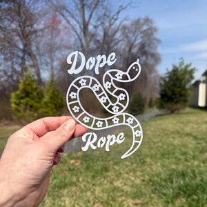 May include: A white vinyl decal featuring a stylised snake with a repeating flower pattern along its body. The text Dope is printed above the snake and the text Rope is printed below the snake in a bold, white, sans-serif font on a clear backing.