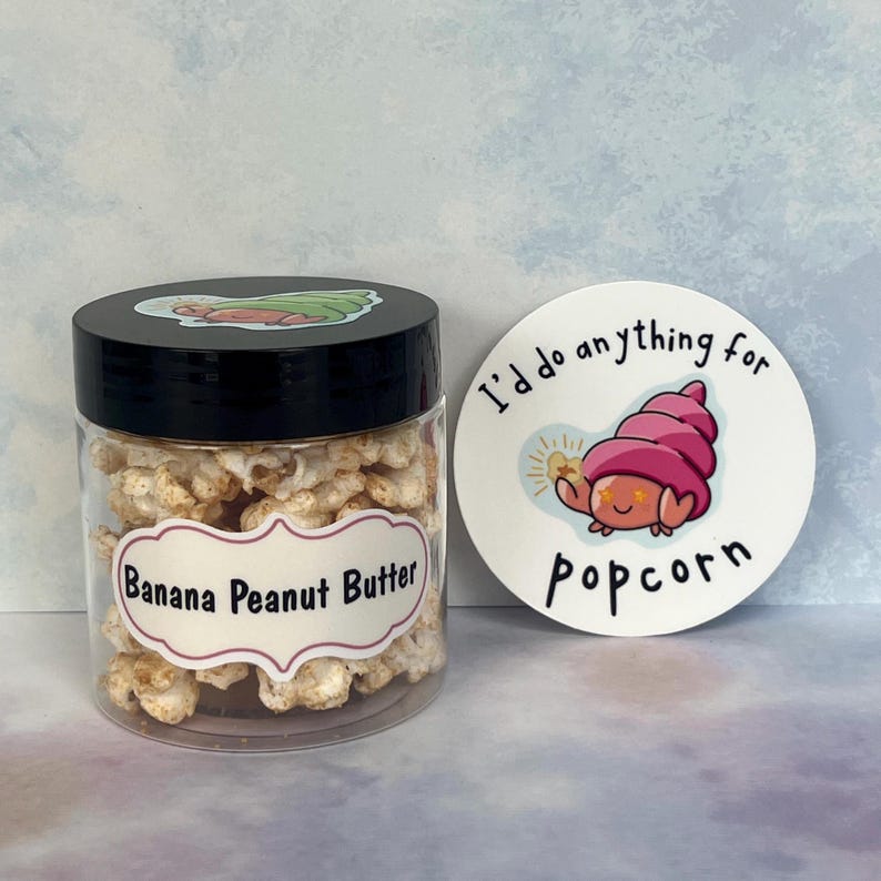 May include: A clear jar of banana peanut butter popcorn with a black lid and a label reading "Banana Peanut Butter". Beside it is a round sticker with the text "I'd do anything for popcorn" and a cartoon character.