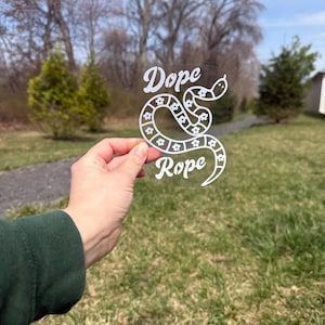 Pode incluir: Um autocolante transparente com as palavras "Dope Rope" numa fonte estilizada. A palavra "Rope" é desenhada como uma cobra com detalhes florais. O autocolante é segurado contra um fundo de relva verde e árvores.