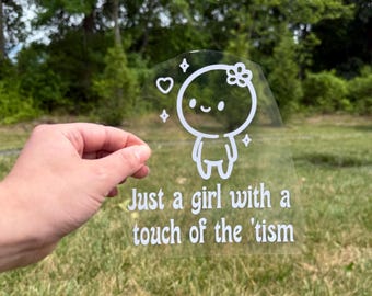 May include: A clear vinyl decal featuring a white line art illustration of a small character with a flower on its head, surrounded by a heart and stars. Below the character, the text reads: Just a girl with a touch of the 'tism.