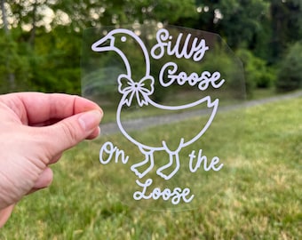 May include: A clear circular decal featuring a white line art illustration of a goose wearing a bow tie. The text "Silly Goose On the Loose" is printed in a white cursive script around the goose illustration on the transparent surface.