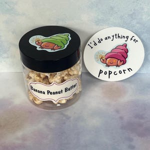 May include: A clear plastic jar filled with banana peanut butter popcorn, with a black lid featuring a cartoon snail. A white sticker next to the jar reads "I'd do anything for popcorn" with a matching snail illustration.