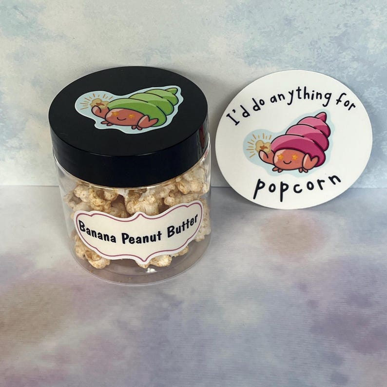 May include: A clear plastic jar filled with banana peanut butter popcorn, with a black lid featuring a cartoon snail. A white sticker next to the jar reads "I'd do anything for popcorn" with a matching snail illustration.