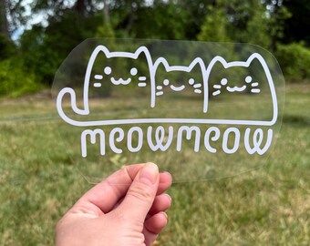 May include: A clear adhesive decal featuring a white line art graphic of three stylised cats sitting side by side. Below the cats, the word meowmeow is written in a rounded, lowercase white font. The design is printed on a transparent backing.