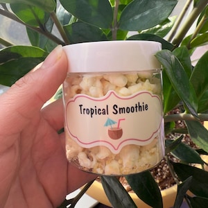 May include: A clear glass jar filled with white popcorn and a label that reads "Tropical Smoothie". The label has a cartoon image of a coconut with an umbrella and a straw.