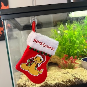 May include: A red Christmas stocking with a white cuff and green and white trim. The stocking has a cartoon snake with a Santa hat holding a gift box. The stocking has the text "Merry Grissmiss" on it.