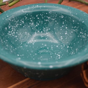 May include: A teal enamel bowl with white speckles. The bowl has a rounded shape and a glossy finish. It is a camping or outdoor dining item.
