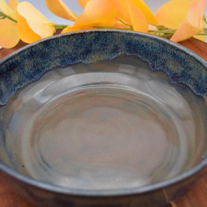 May include: A handmade ceramic bowl with a wide, shallow design. The bowl features a dark blue rim with a textured edge, transitioning to a light brown and gray interior. The bowl is displayed on a wooden surface, with yellow flowers in the background.