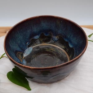 May include: A handmade ceramic bowl with a dark brown exterior and a deep blue interior. The bowl's rim has a textured, wavy design. The interior features a swirling pattern of blues, greens, and browns. The bowl is set on a white cloth.