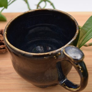 May include: A handmade ceramic mug with a dark blue interior and a brown exterior. The mug has a unique handle and a slightly irregular shape. The rim and handle are accented with gold-colored details. The mug is placed on a wooden surface.