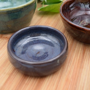 May include: A collection of three handmade ceramic bowls in varying shades of blue, green, and brown. The bowls are glazed and have a glossy finish. They are arranged on a light brown bamboo surface.