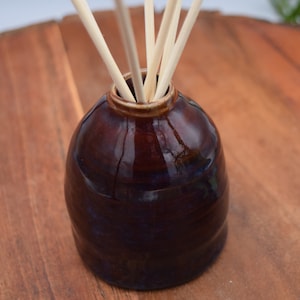 May include: A dark brown ceramic essential oil diffuser with a glossy finish. The diffuser has a rounded shape and a narrow opening, with several light-colored reeds inserted. The diffuser sits on a wooden surface.