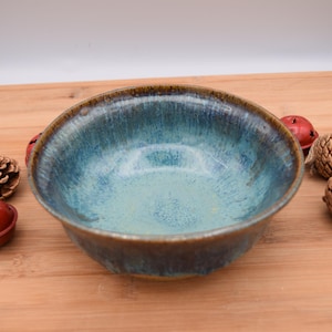 May include: A handmade ceramic bowl with a teal and brown glaze. The bowl is round and has a smooth interior. Decorative pinecones and red bells surround the bowl on a wooden surface.