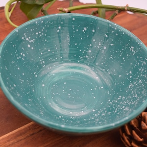 May include: A teal ceramic bowl with white speckles. The bowl is round with a slightly angled rim. The background includes a wooden surface, green leaves, and a pine cone. This bowl is suitable for serving food.