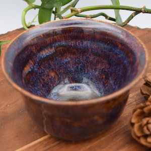 May include: A handmade ceramic bowl with a rich, glazed interior featuring shades of deep purple and blue. The exterior has a warm brown tone. The bowl sits on a wooden surface, with greenery and pine cones in the background.