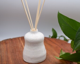 May include: A white ceramic essential oil diffuser with a speckled design, holding several wooden reeds. The diffuser sits on a wooden surface, with green leaves in the background. The ceramic vessel has a rounded shape.