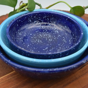 May include: Three nested ceramic bowls in shades of blue, with a speckled white pattern. The bowls are arranged on a wooden surface, with a plant in the background. The bowls are likely for serving or food preparation.