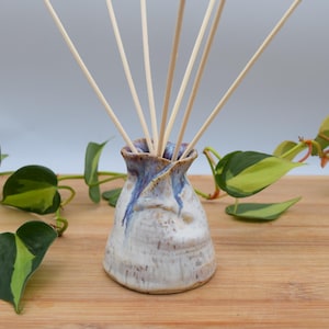 May include: A handmade ceramic essential oil diffuser with a white and blue glaze. The diffuser holds several wooden reeds and is surrounded by green and yellow heart-shaped leaves. The diffuser sits on a wooden surface.