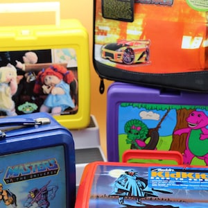 May include: A collection of five rectangular lunch boxes featuring various character graphics including Cabbage Patch Kids, Masters of the Universe, Barney, and a race car. The lunch boxes are yellow, blue, purple, red, and black with plastic handles.