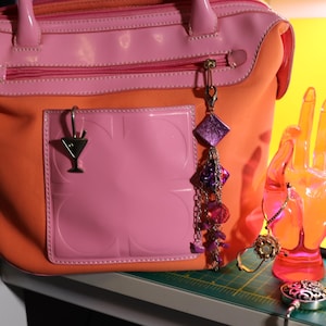 May include: A two-tone handbag in orange and pink with a pink pocket and a cocktail charm. A decorative hand and other charms are next to the bag. The bag has a top handle and a zipper.