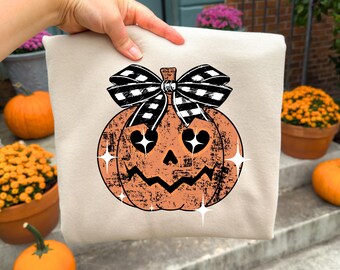 May include: A beige sweatshirt with a distressed orange jack-o'-lantern design and a black and white checkered bow. The pumpkin has star-shaped eyes and a jagged mouth. The sweatshirt is held up by a hand.
