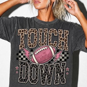 Könnte beinhalten: Ein anthrazitfarbenes T-Shirt mit einem Grafikdruck, der das Wort TOUCH in Leopardenmuster, einen rosa Football und das Wort DOWN in Leopardenmuster zeigt. Das Design enthält einen schwarz-weißen Schachbrett-Hintergrund, rosa Blitze und Sterne.